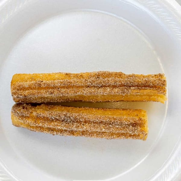 Two golden-brown churros, coated in cinnamon sugar, rest on a white disposable plate.